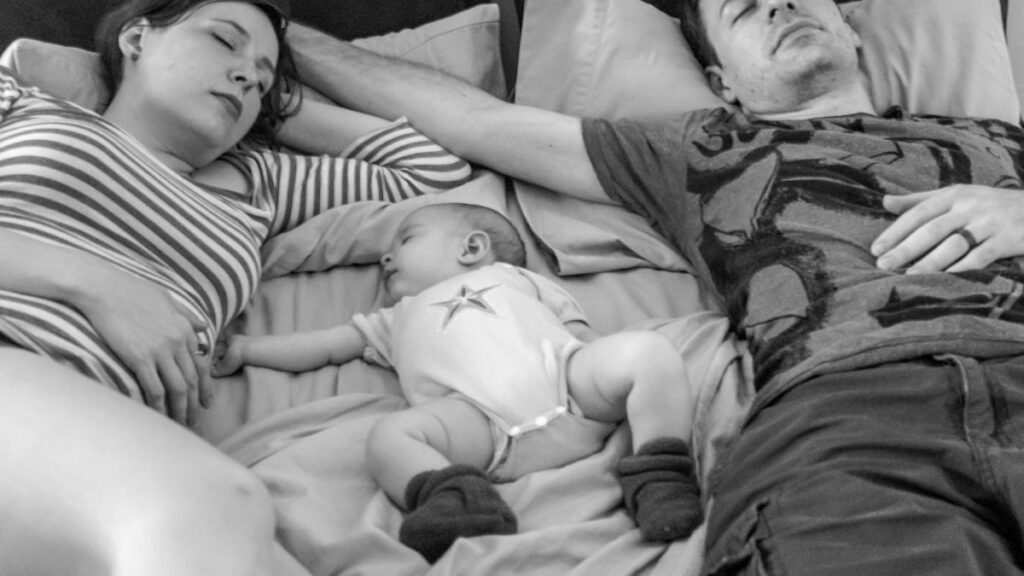 A couple lying in bed with their newborn, tender and tired — marriage after baby