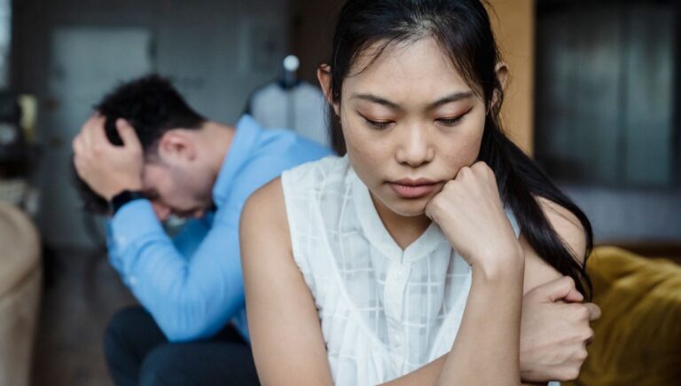 Woman looking hurt and withdrawn while her frustrated partner sits behind her after a misread comment