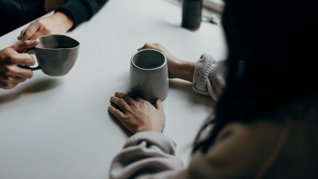 Two people holding coffee mugs at a table, one of the simplest rituals of connection
