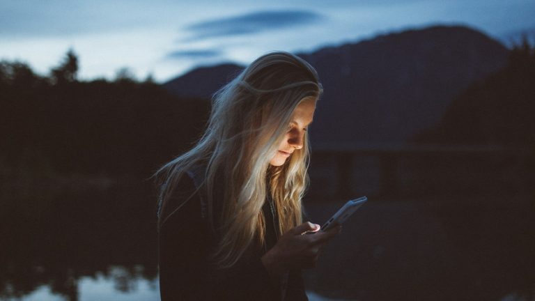 Woman reading messages on her phone by the water, the kind of quiet moment romance scams exploit